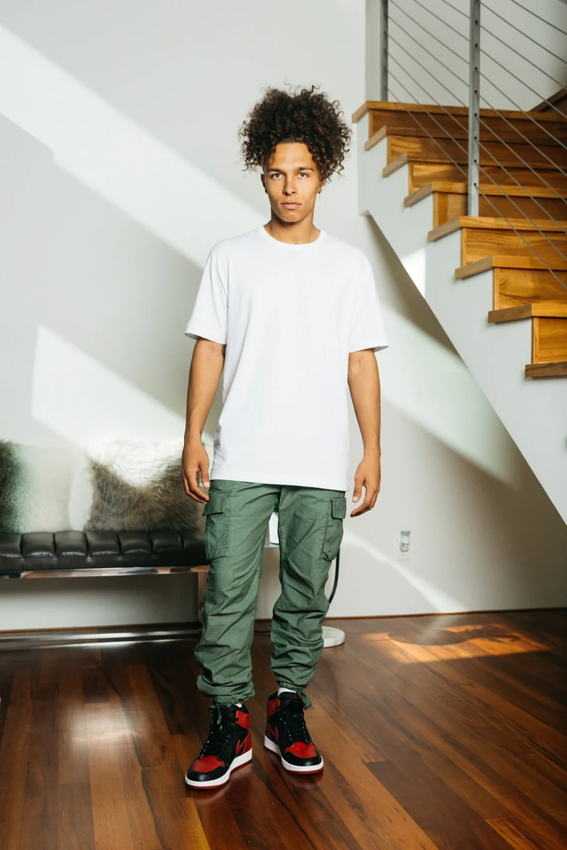 Full-length portrait in a white tee and green trousers.