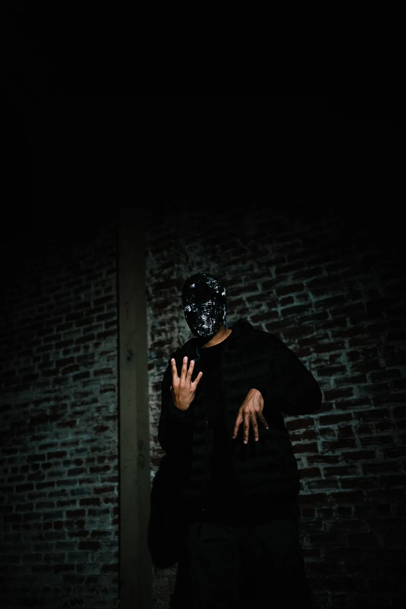 Masked WRKINSILENCE portrait against a dark brick wall.
