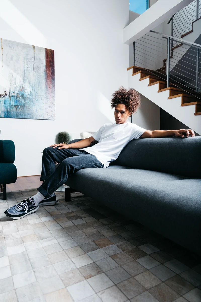 Model in a white tee reclining on a couch in a bright living room.