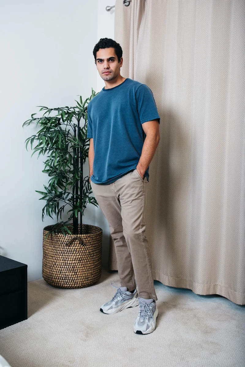 Standing portrait in a blue tee beside a plant and pale curtains.