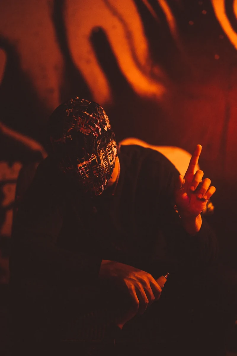WRKINSILENCE seated in a mask under orange light.