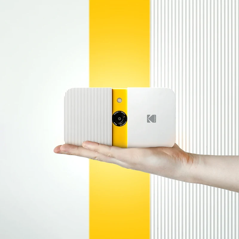 Hand holding a white Kodak camera against a yellow stripe.