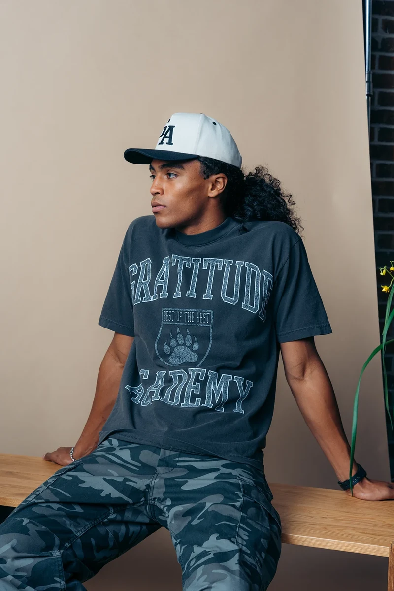 Seated model wearing a dark Gratitude Academy tee and camouflage trousers.
