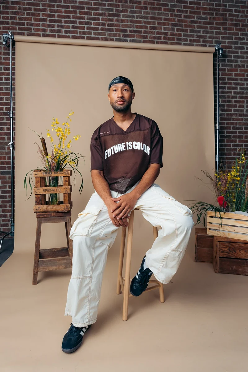 Seated portrait in a brown graphic tee and cream trousers.