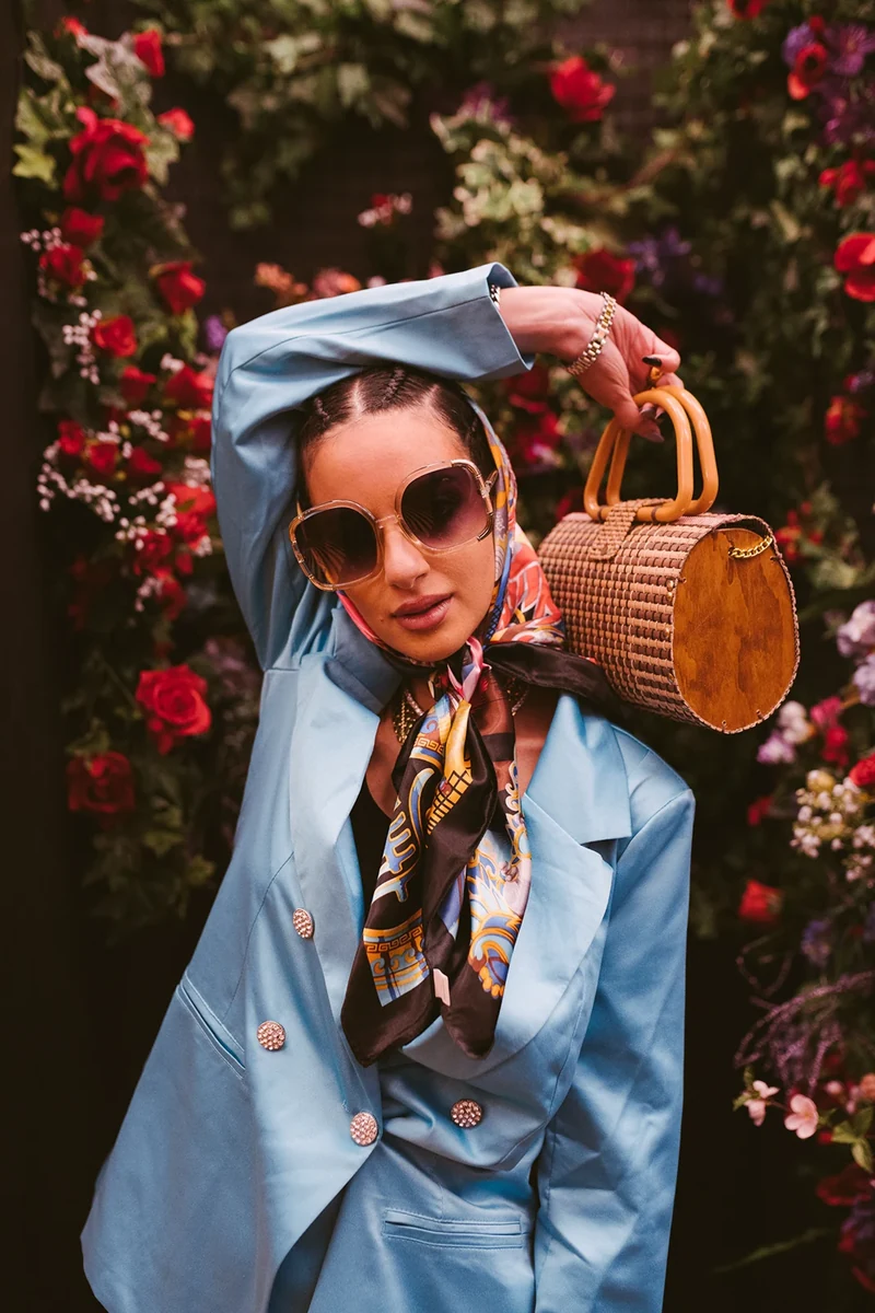 ZEOPATRA in a blue suit with a round woven handbag against flowers.