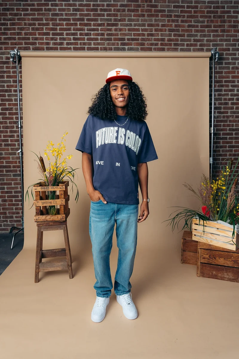 Full-length portrait in a navy graphic tee, jeans, and a baseball cap.