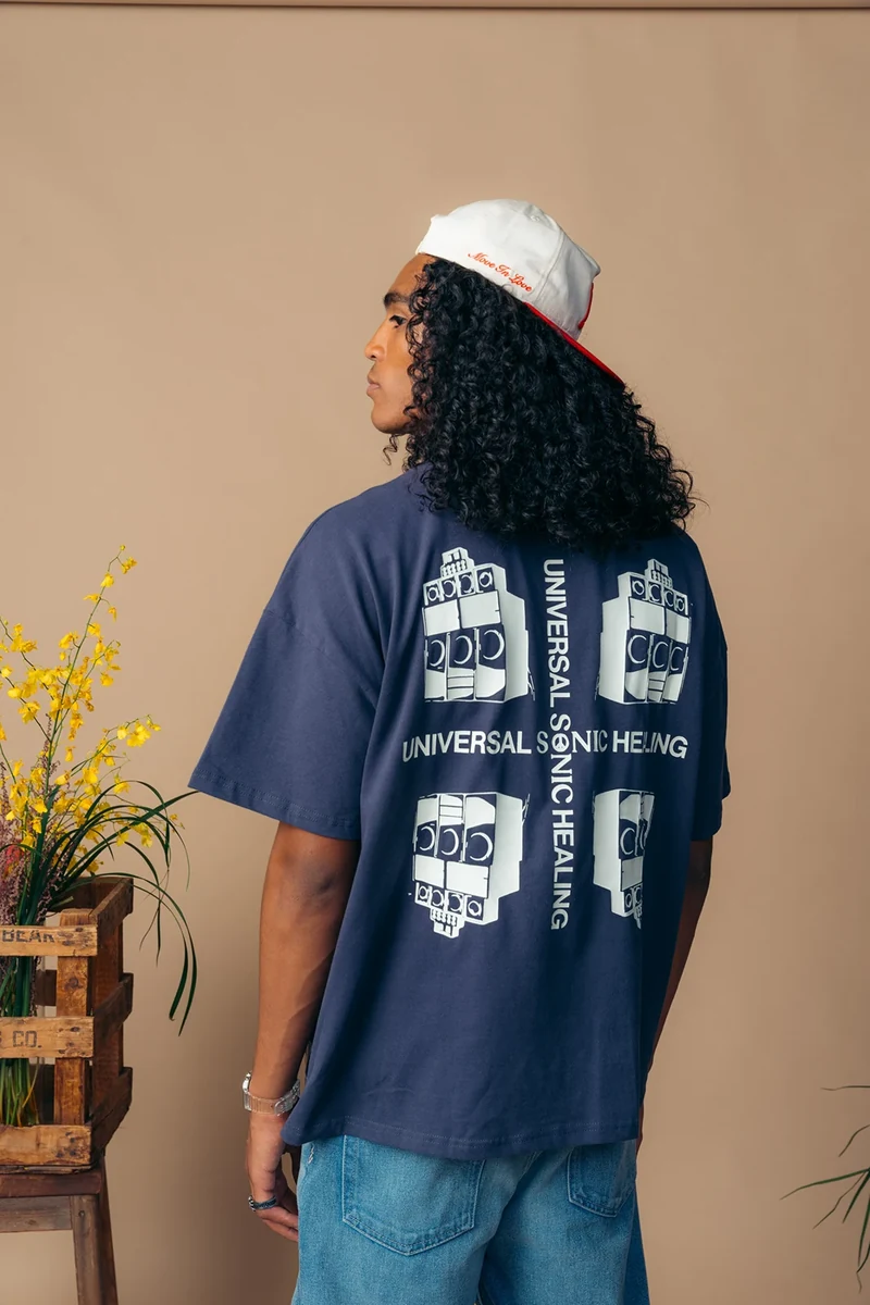 Back print on a navy graphic tee, photographed beside yellow flowers.
