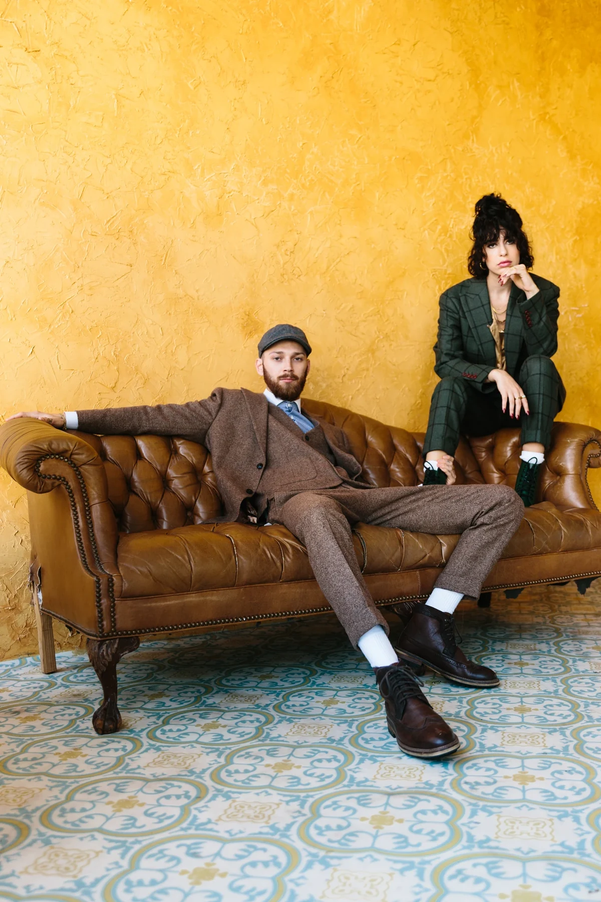 Two models in relaxed tailoring on a leather couch against a yellow wall.