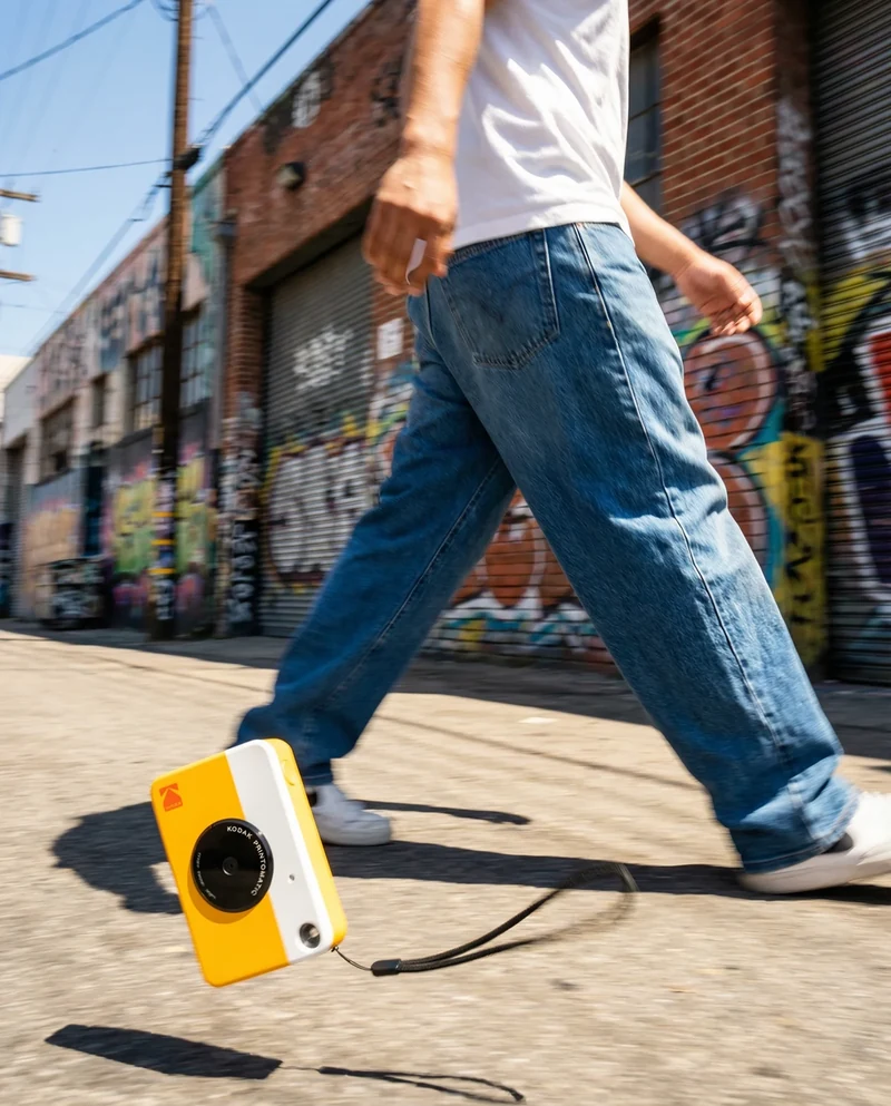 Yellow Kodak camera swinging from a strap in a street scene.