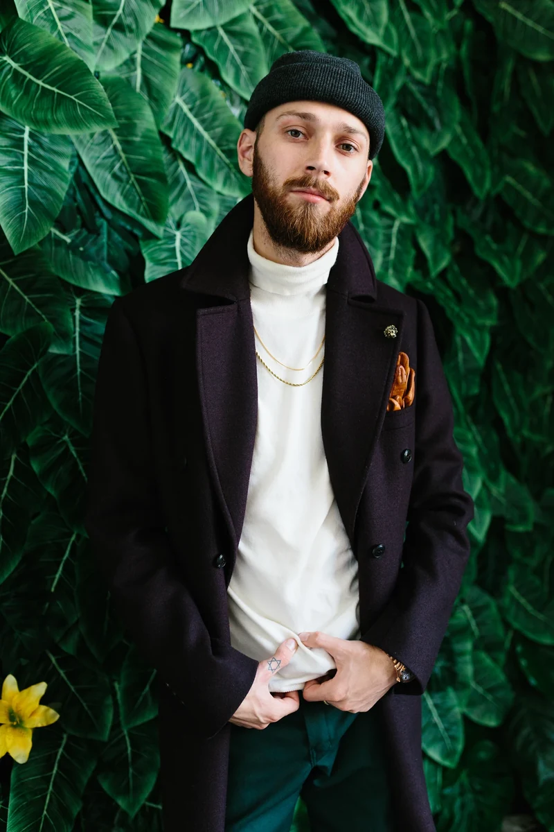 Portrait of a model in a dark blazer and knit hat against foliage.