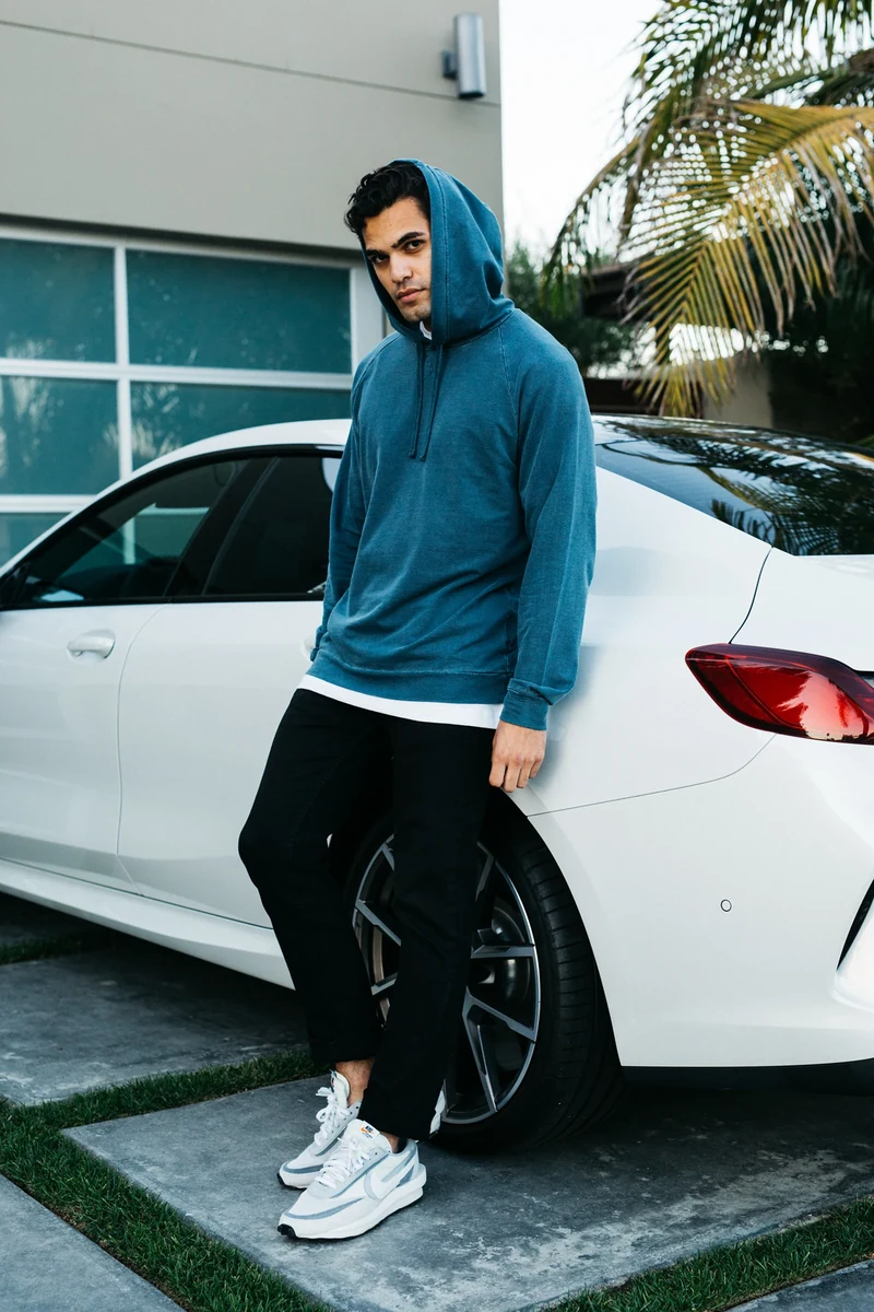 Model in a blue hoodie leaning against a white car.